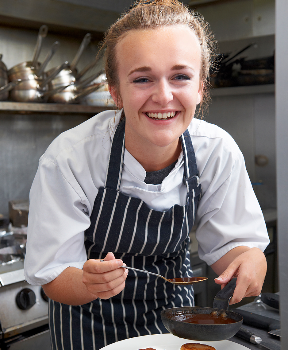 Young adult female working in a kitchen and pouring sauce on plate