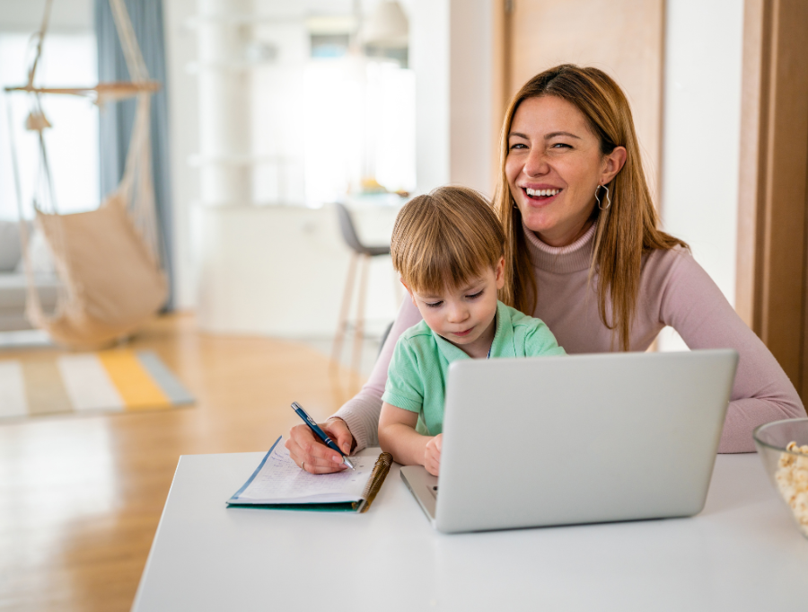 Woman with long brown hair sits with a young boy in her lap with a laptop in front of them. She writes on a notepad.